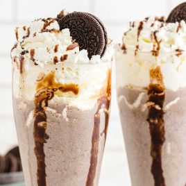 two Oreo Milkshake in a tall glass topped with whip cream and Oreos in a bowl